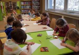 Dzieci w bibliotece siedzą przy stolikach i rysują na białych kartkach